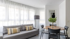 A modern living room with a grey sofa, two cushions, a round wooden table with four chairs, a tall floor lamp, and a potted plant.