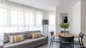 Una sala moderna con un sofá gris, dos cojines, una mesa redonda de madera con cuatro sillas, una lámpara de pie alta y una planta en maceta.