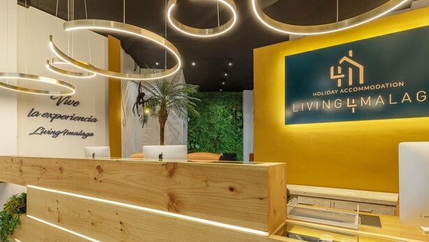 Un área de recepción con un mostrador de madera, una pared con un logotipo y texto, y una pared verde con plantas.