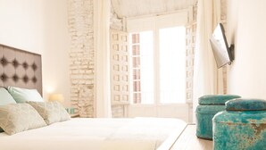 A bedroom with a large bed, a stone wall, a window with shutters, and a TV mounted on the wall.