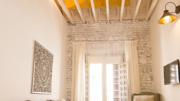 A cozy room with a stone wall, a wooden ceiling, a large window with shutters, a sofa, a chair, and a wall-mounted light fixture.