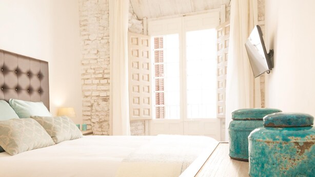 A bedroom with a bed, a stone wall, a window with shutters, and a TV mounted on the wall.