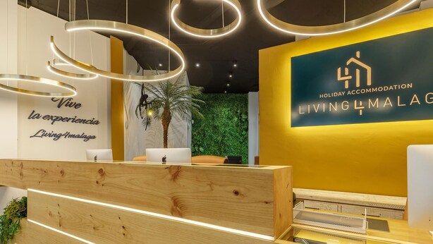 A reception area with a wooden desk, a wall with a green wall and a palm tree, and a sign for 'Holiday Accommodation Living4Malaga'. - Mariblanca 1D Superior Living4malaga (Málaga)