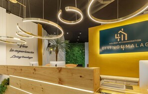Una zona de recepción con un escritorio de madera, una pared decorada con vegetación verde y una palmera, y un cartel que indica 'Holiday Accommodation Living4Malaga'.