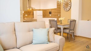A modern living room with a beige sofa, a dining table with chairs, a kitchen in the background, and a wall clock. - Mariblanca 1D Superior Living4malaga (Málaga)