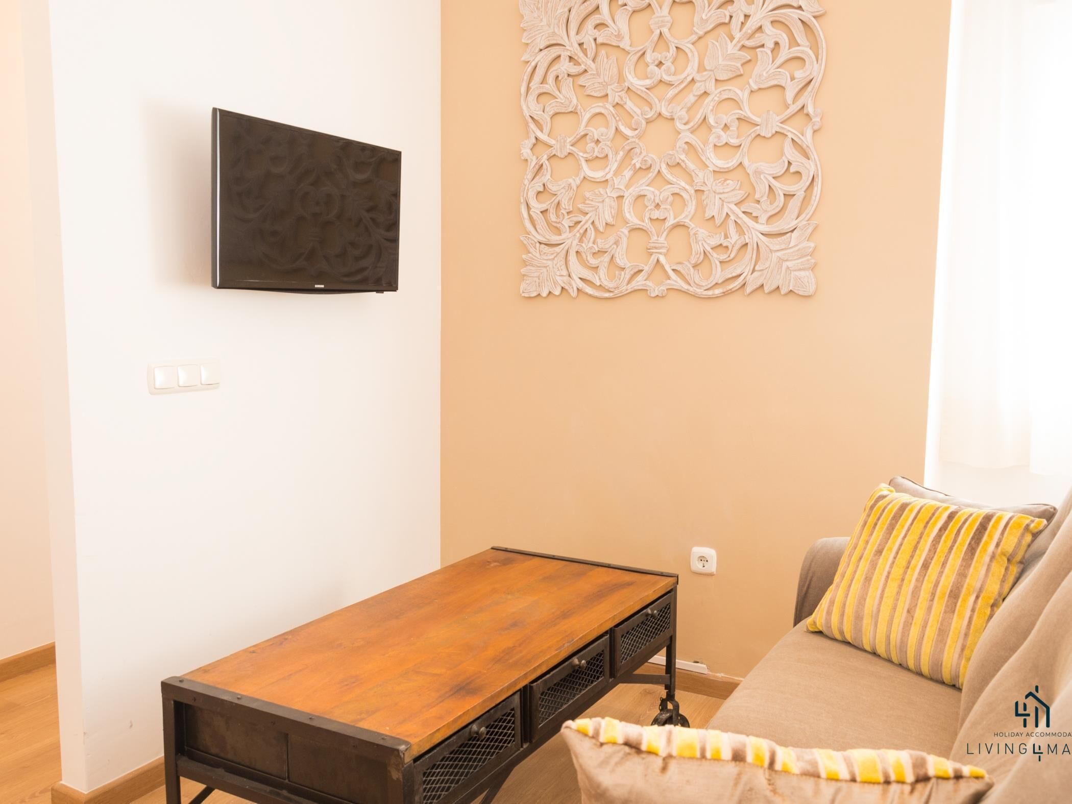 A living room with a wooden coffee table, a sofa with striped cushions, a wall-mounted TV, and a decorative wall panel.