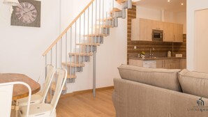 A modern apartment with a wooden staircase, a dining area with chairs, a sofa, and a kitchen with wooden cabinets and a microwave.