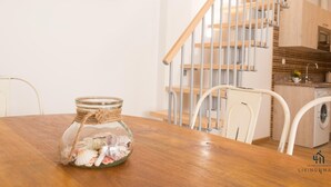 A wooden dining table with a glass jar containing seashells, a rustic wall clock, and a staircase with metal railings.