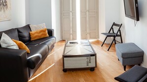 A modern living room with a black leather sofa, a coffee table, a grey ottoman, and a TV mounted on the wall.