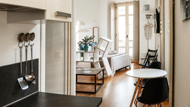A modern kitchen with a dining area, a clock on the wall, and a view into the living room.