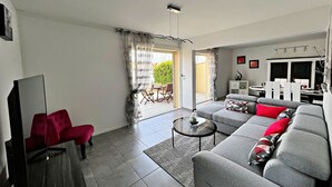 Ein modernes Wohnzimmer mit grauem Sofa, rotem Sessel und einem kleinen Couchtisch. Es gibt einen Essbereich mit weißem Tisch und Stühlen. Das Zimmer verfügt über ein großes Fenster mit Vorhängen und eine Tür, die zu einem Außenbereich führt.