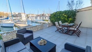 Eine Terrasse mit Korbmöbeln, einem Tisch mit Tablett und Blick auf einen Yachthafen mit Booten.