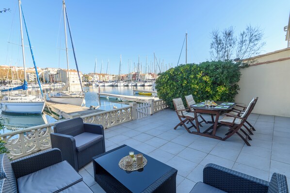 Terraço com móveis de vime, uma mesa com bandeja e vista para uma marina com barcos.