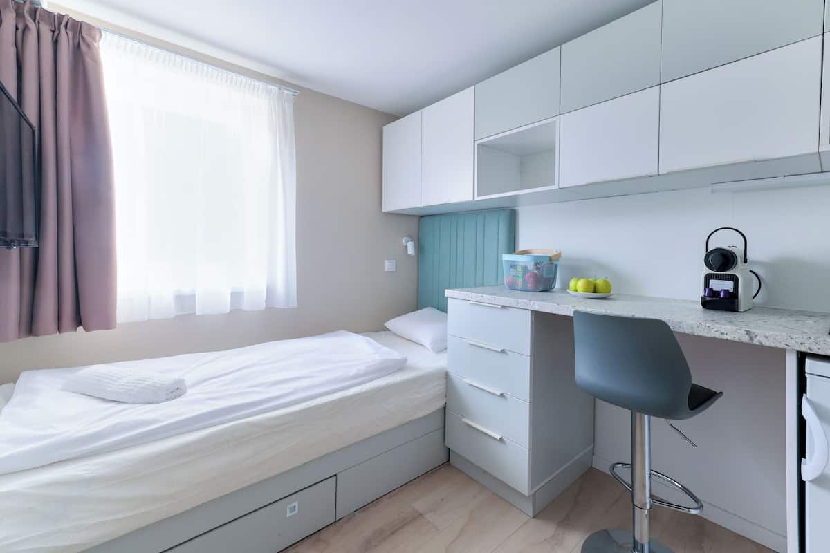 A compact room with a bed, a kitchenette, a bar stool, and a window with curtains.