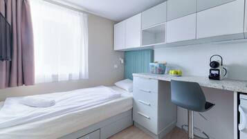 A compact room with a bed, a kitchenette, a bar stool, and a window with curtains.