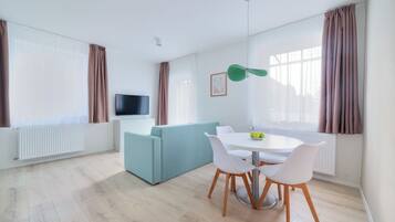 A modern living room with a sofa, a dining table with chairs, a flat-screen TV, and a window with curtains.
