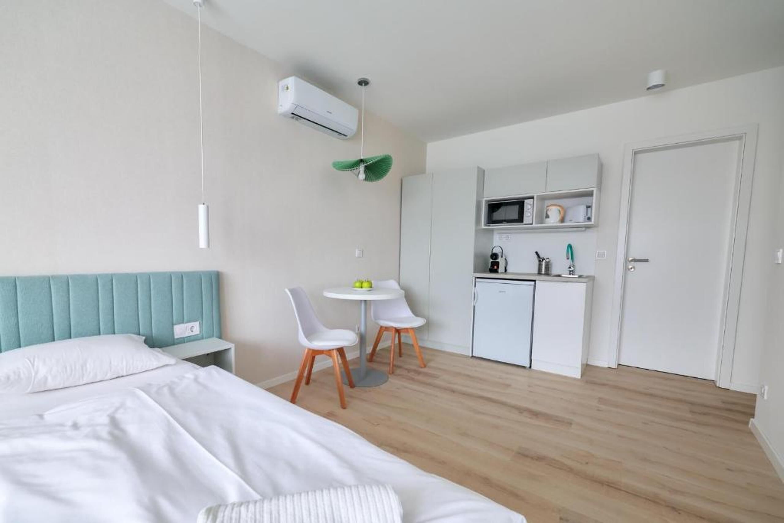 A modern hotel room with a bed, a small kitchenette, a dining area with two chairs, and a ceiling fan.