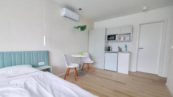 A modern hotel room with a bed, a small kitchenette, a dining area with two chairs, and a ceiling fan.