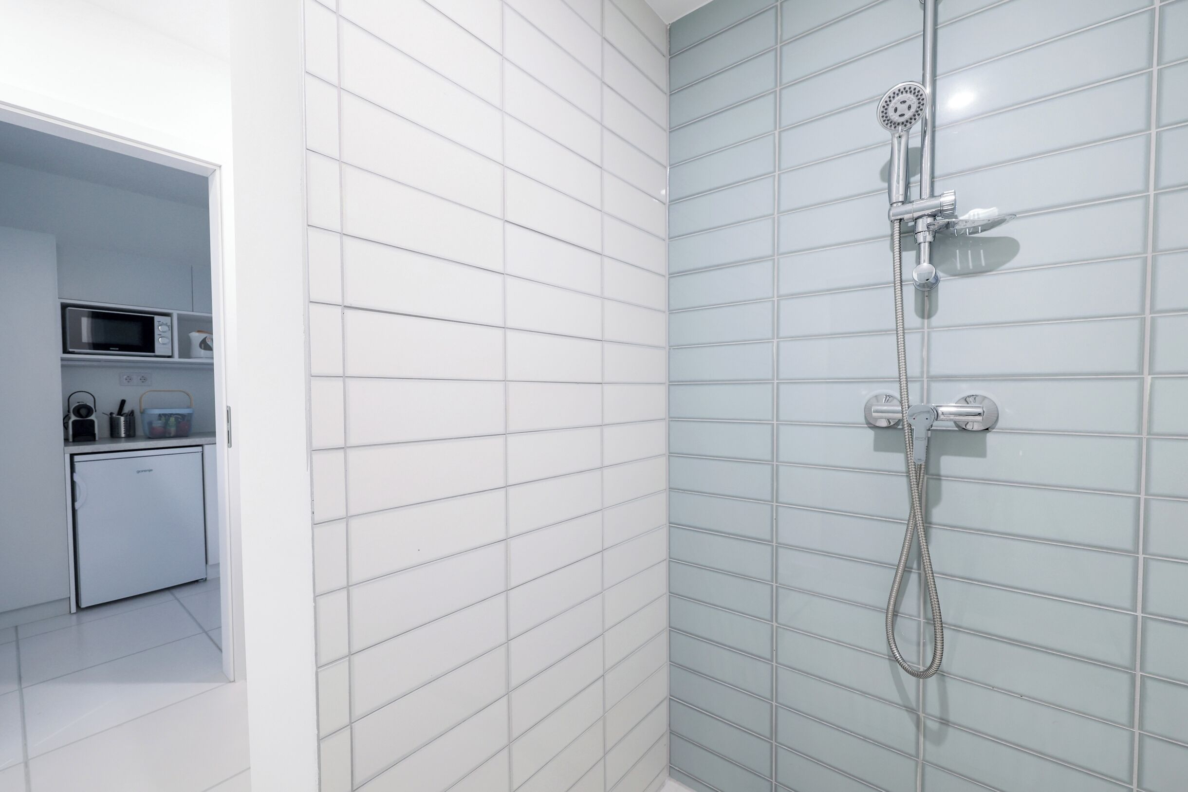 Een moderne badkamer met wandtegels in het wit en lichtblauw, een douche met een handdouche, en een klein keukentje met een magnetron en een koelkast.