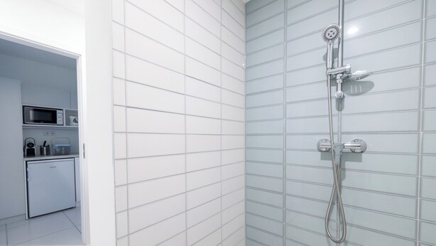 Un baño moderno con paredes de azulejos blancos y azul claro, una ducha con cabezal de ducha manual y una pequeña área de cocina con microondas y refrigerador.