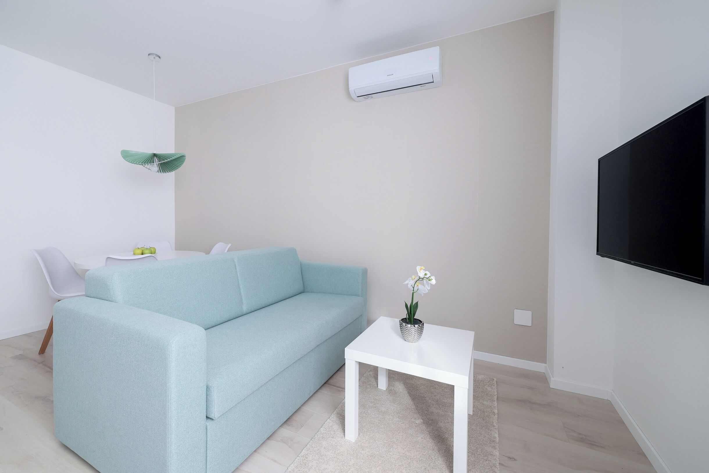A modern living room with a light blue sofa, a white coffee table with a potted orchid, a flat-screen TV mounted on the wall, and a ceiling fan.
