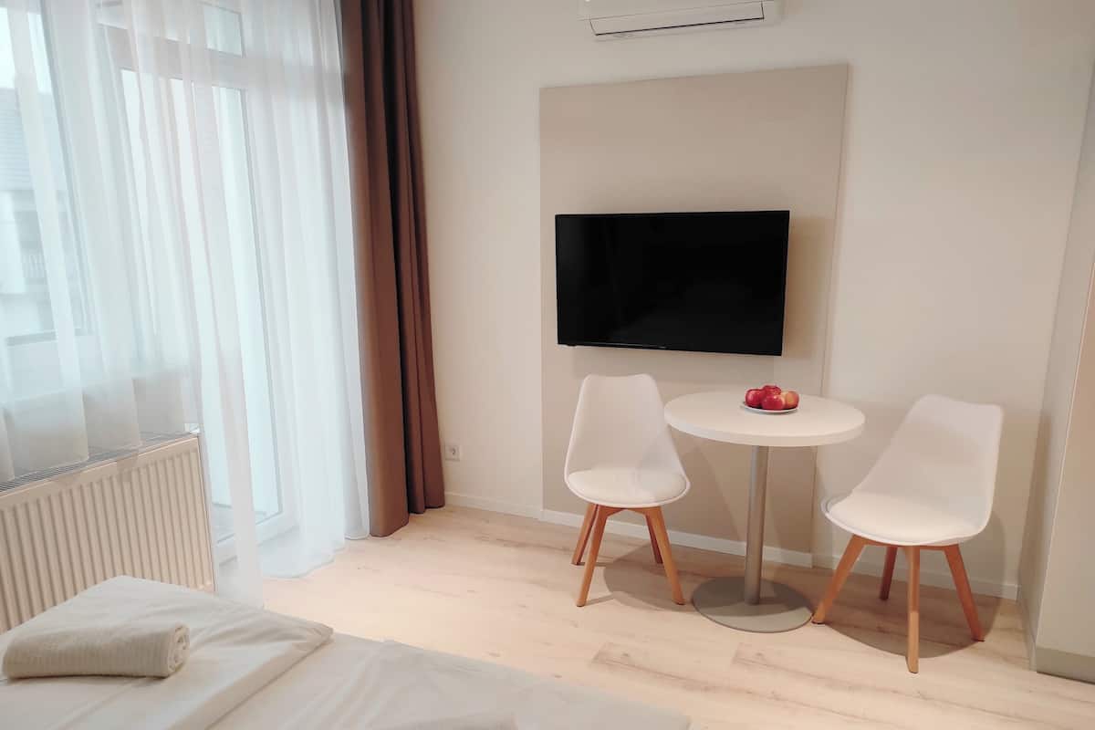A hotel room with a bed, a TV, two chairs, a small table with apples, and a window with curtains.