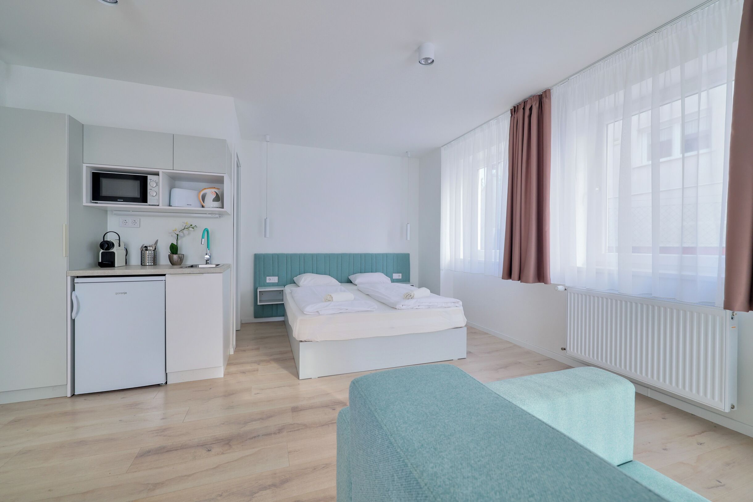 A modern hotel room with a kitchenette, a bed with white bedding, a teal bench, and large windows with curtains.