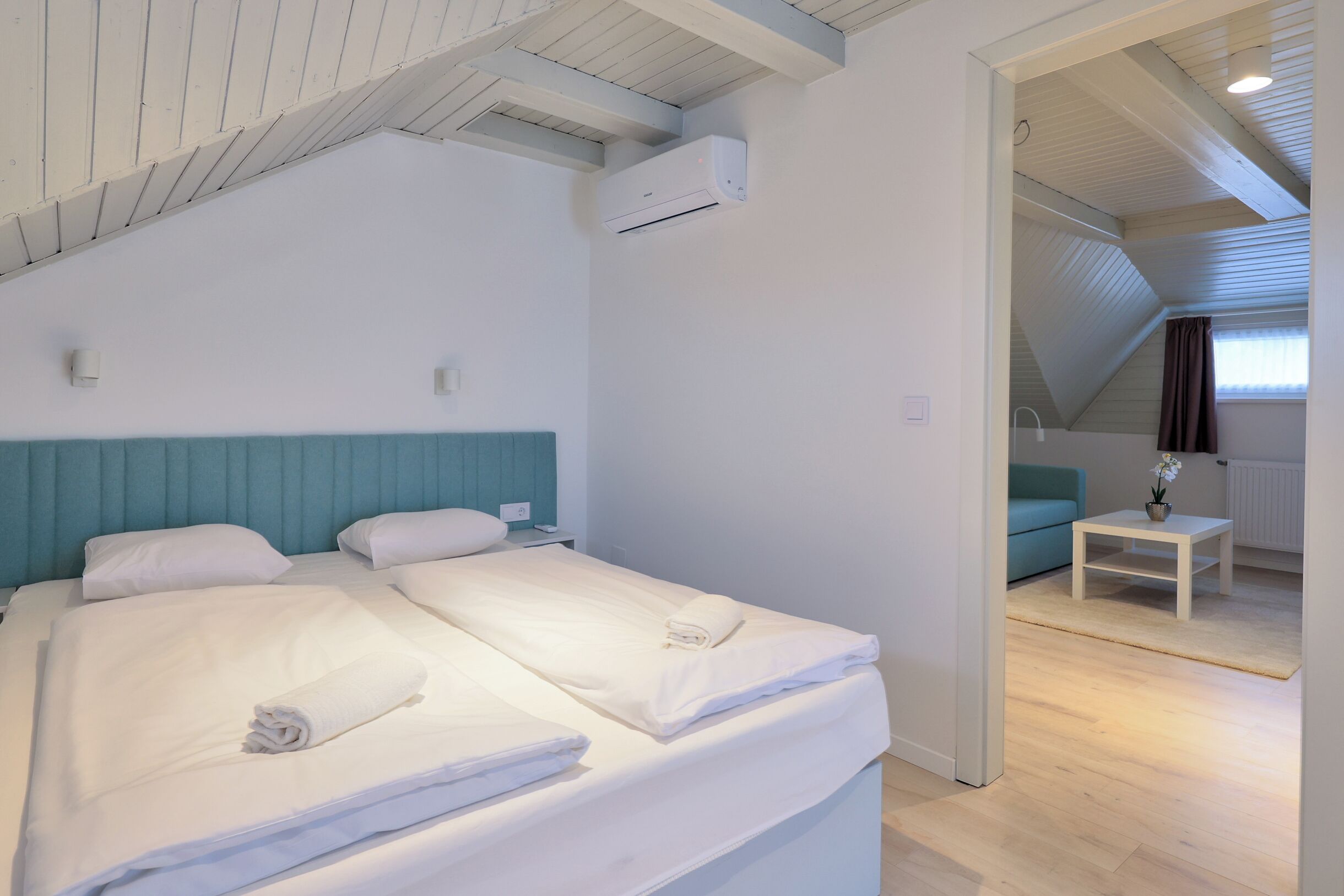 Ein Schlafzimmer mit zwei Betten, einem blauen Kopfteil, einem kleinen Tisch mit Pflanze und einem Fenster mit Vorhängen.