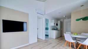 Cocina moderna con armarios blancos, zona de comedor con una mesa redonda y sillas blancas, y un televisor de pantalla plana montado en la pared.