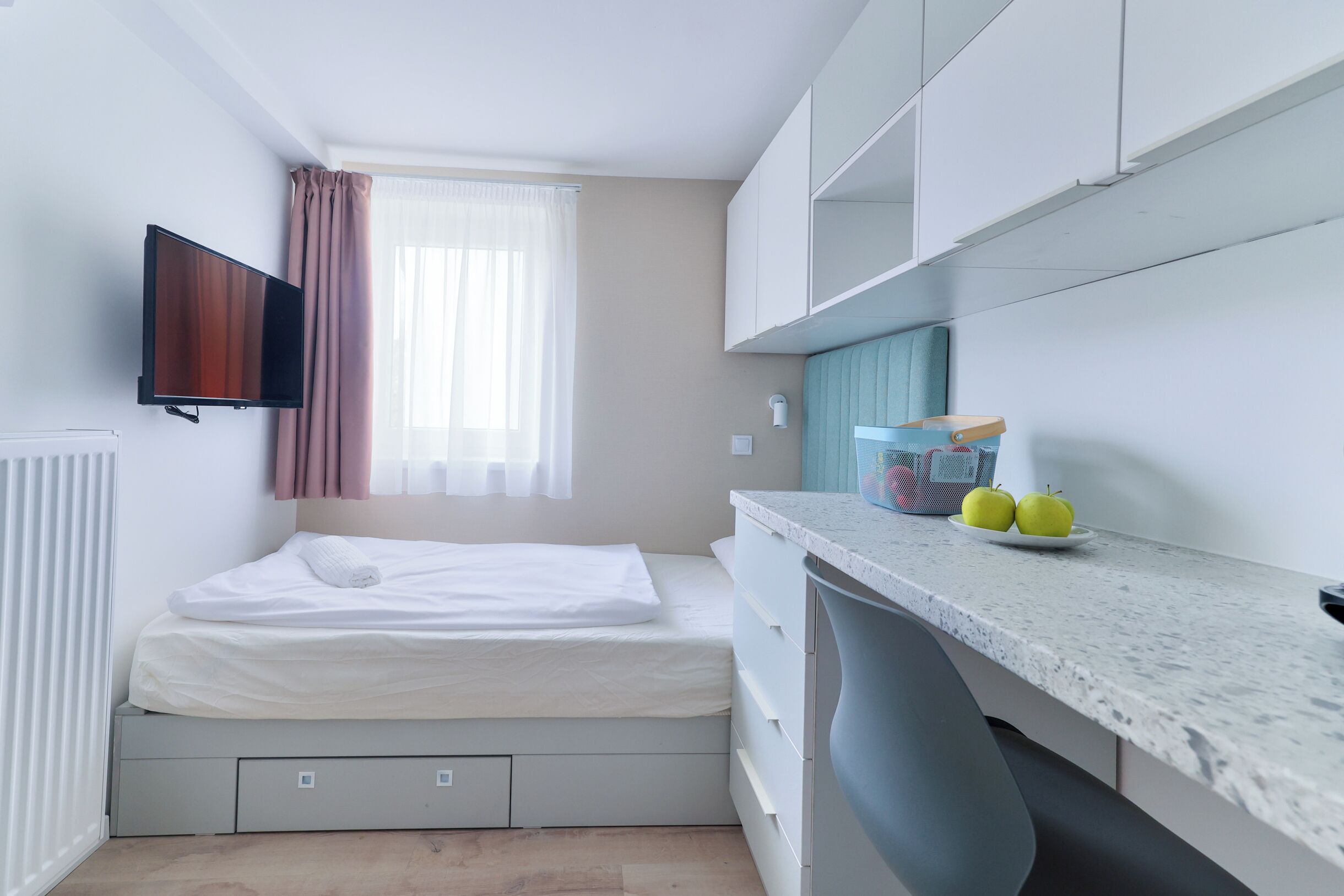 A compact room with a bed, a TV mounted on the wall, a window with curtains, a kitchenette area with a countertop and a fruit bowl.