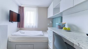 A compact room with a bed, a TV mounted on the wall, a window with curtains, a kitchenette area with a countertop and a fruit bowl.