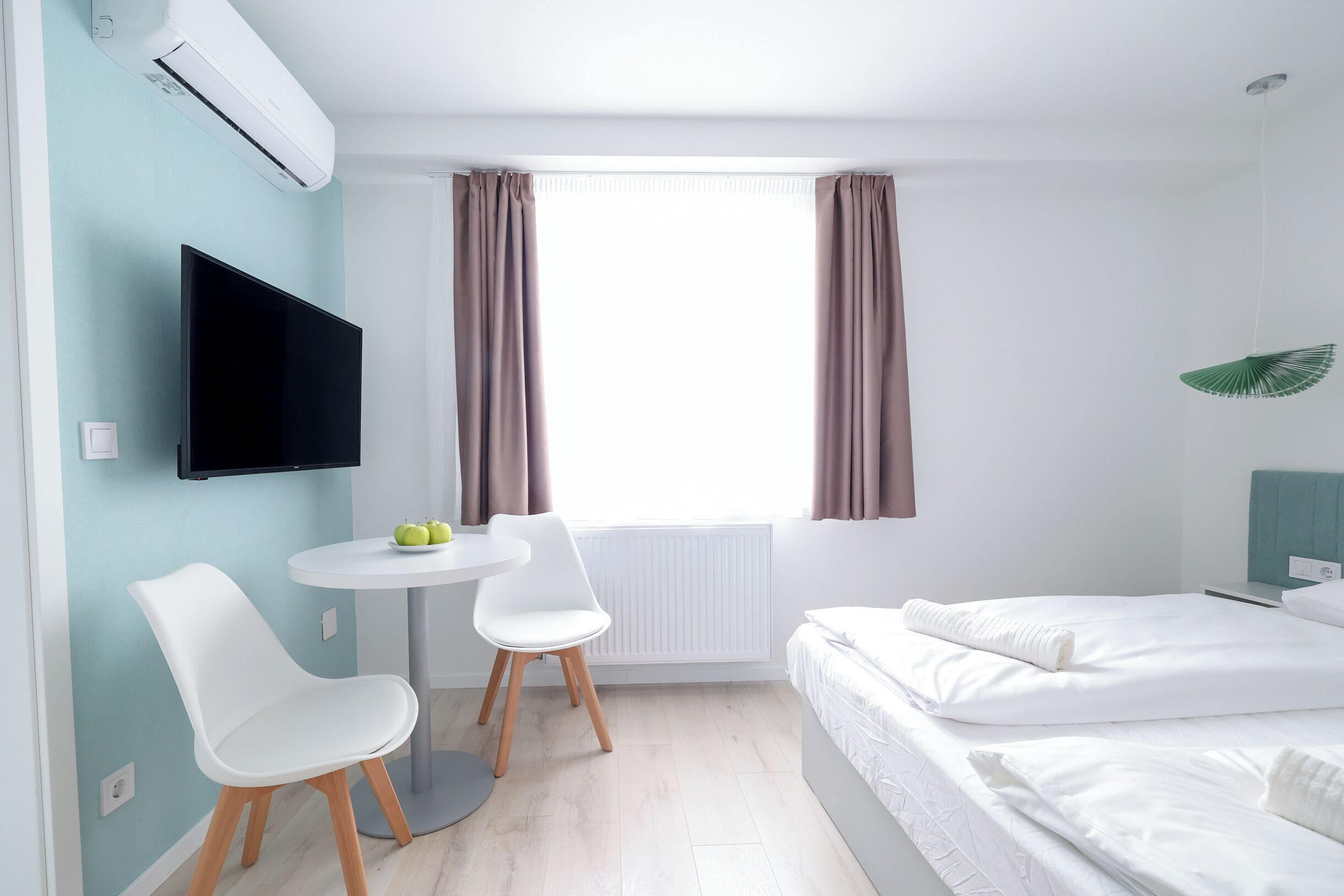 A modern hotel room with a bed, a TV, a small table with chairs, and a window with curtains.