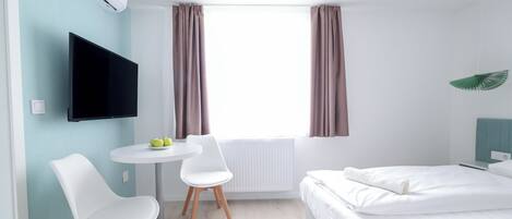 Una habitación de hotel moderna con cama, televisor, una mesita con sillas y un ventanal con cortinas.