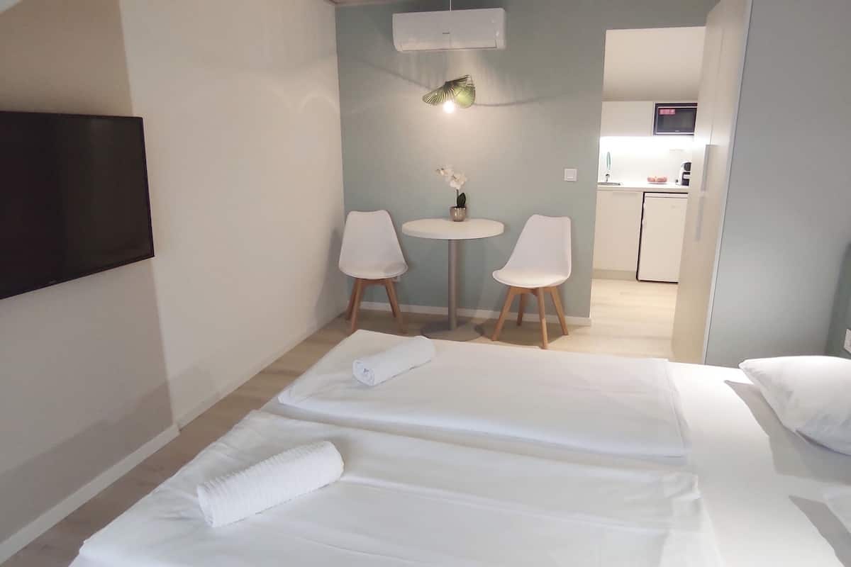 A hotel room with two beds, a TV mounted on the wall, a small table with chairs, and a kitchenette in the background.