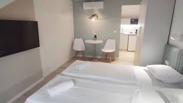 Ein Hotelzimmer mit zwei Betten, einem an der Wand befestigten Fernseher, einem kleinen Tisch mit Stühlen und einer Küchenzeile im Hintergrund.