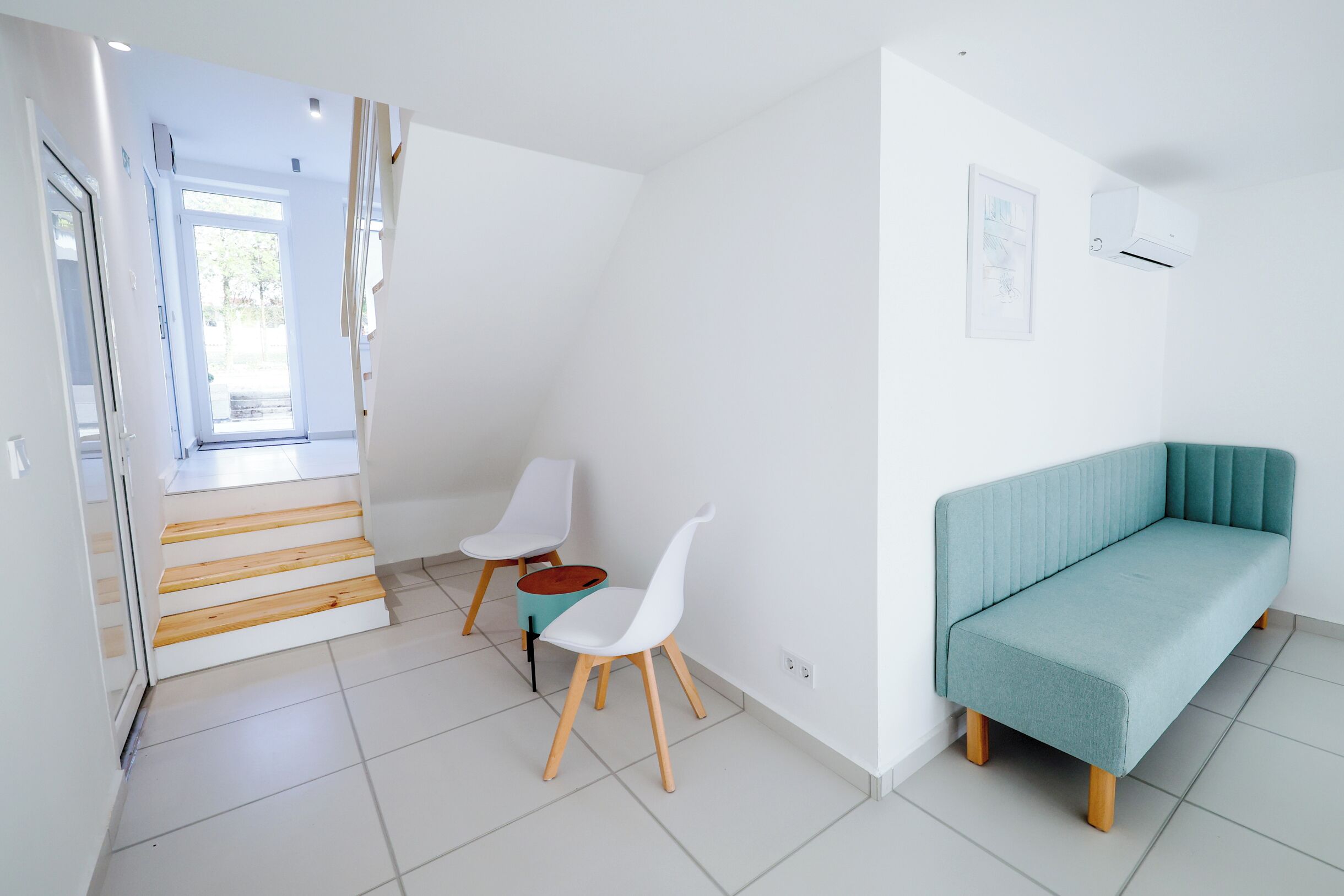 A modern living room with a teal sofa, white chairs, and a staircase leading to another room.