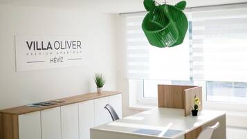 Um espaço de escritório moderno e minimalista com uma mesa branca, uma cadeira e uma luminária pendente verde. Uma placa na parede diz "VILLA OLIVER PREMIUM APARTMAN HÉVIZ".