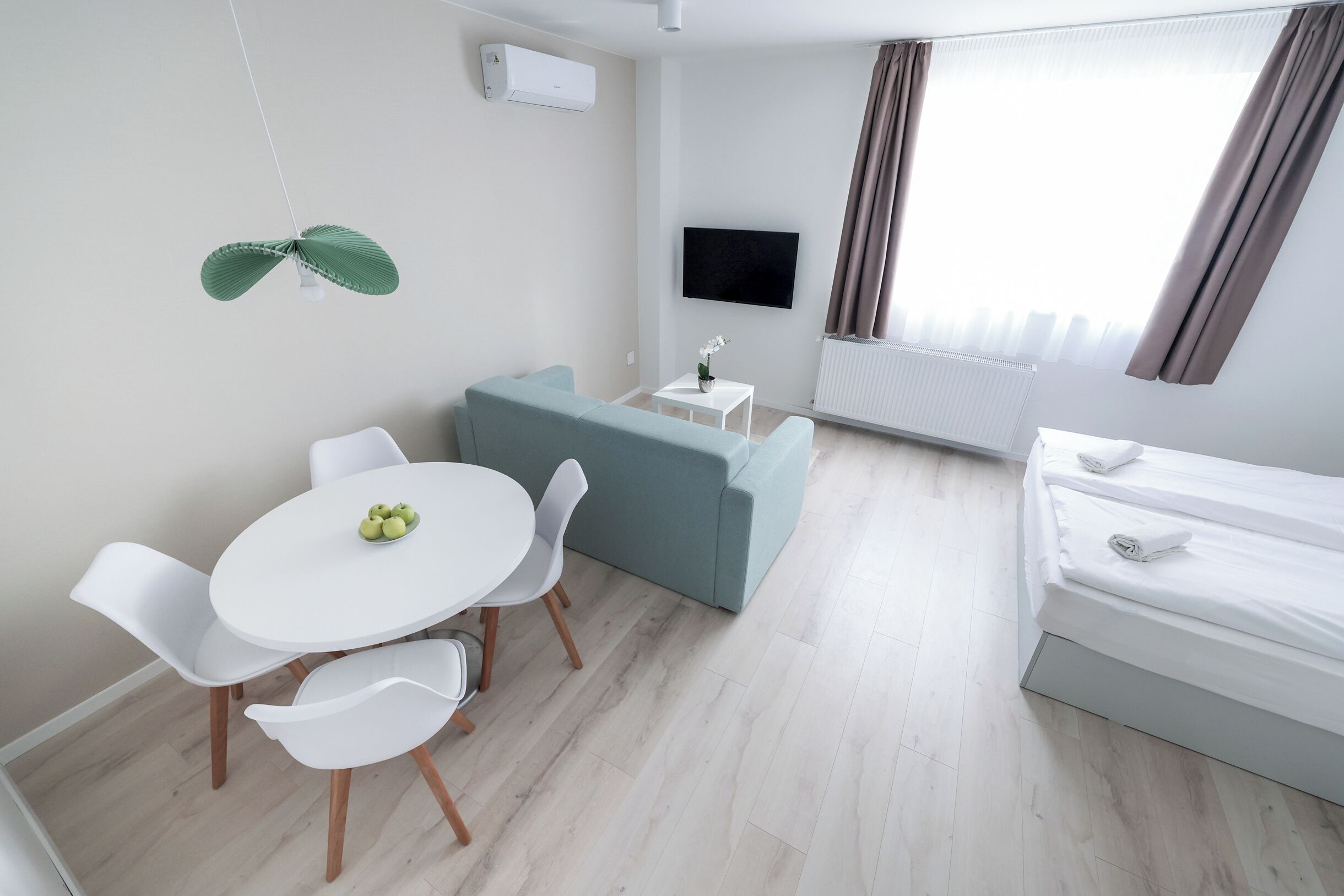 A modern hotel room with a sofa, a dining table with chairs, a TV mounted on the wall, and a bed with neatly made sheets.
