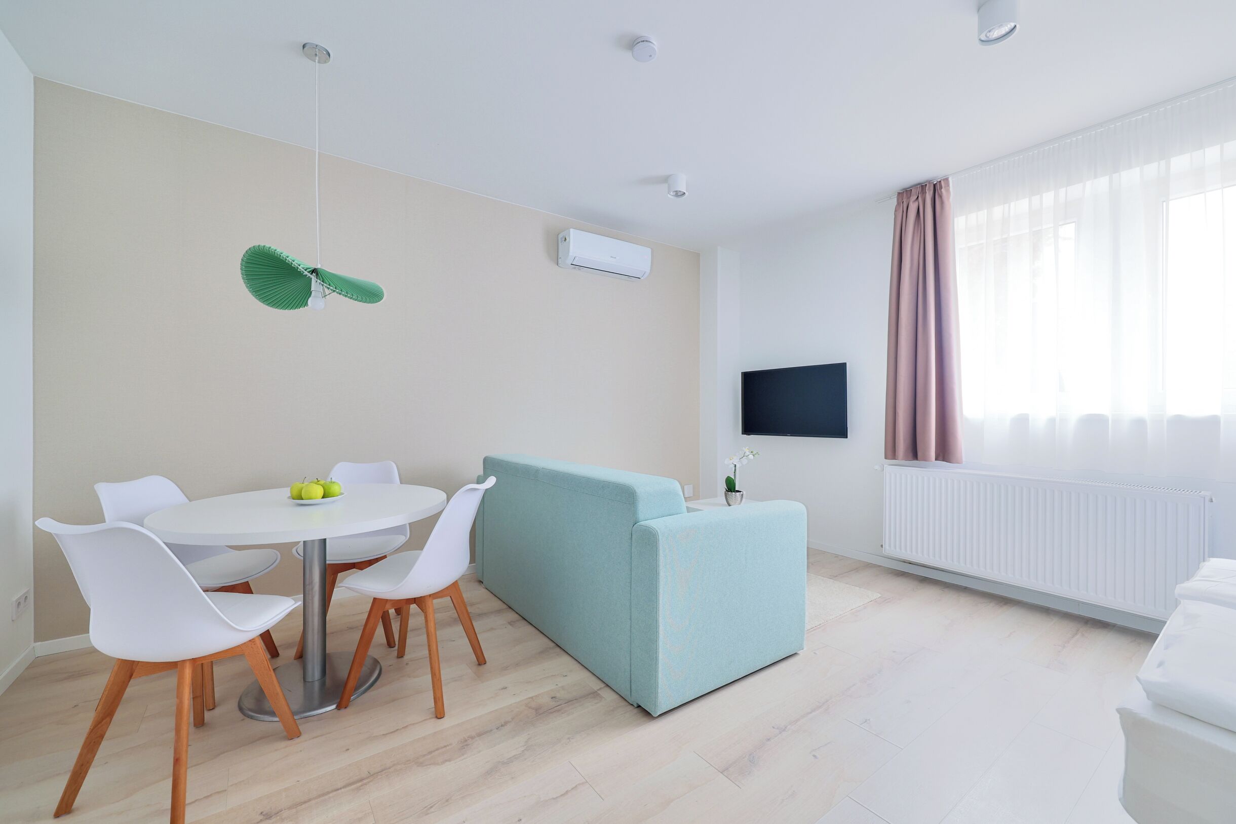 A modern living room with a light blue sofa, a round dining table with white chairs, and a flat-screen TV mounted on the wall.