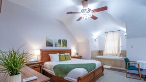 A bedroom with a wooden bed, a nightstand, a bedside table with a plant, a ceiling fan, and a bathroom with a bathtub and shower curtain. - Mountain Meadows Retreat - A Romantic Escape Overlooking the Ozark Mountains (Reeds Spring)