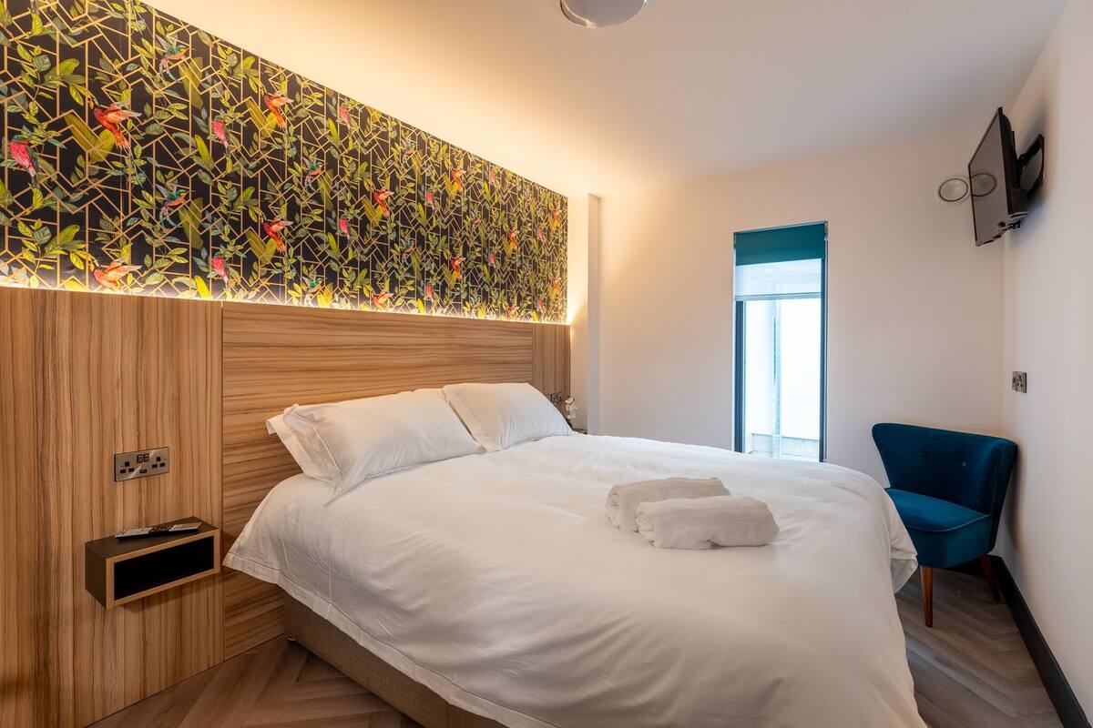 A modern hotel room with a large bed, a wooden headboard, a wall-mounted TV, and a blue chair.