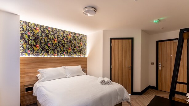 A modern hotel room with a wooden headboard, a large window with a geometric patterned view, a neatly made bed with white linens, and a wooden floor. - Hotel No.9 (Londonderry)