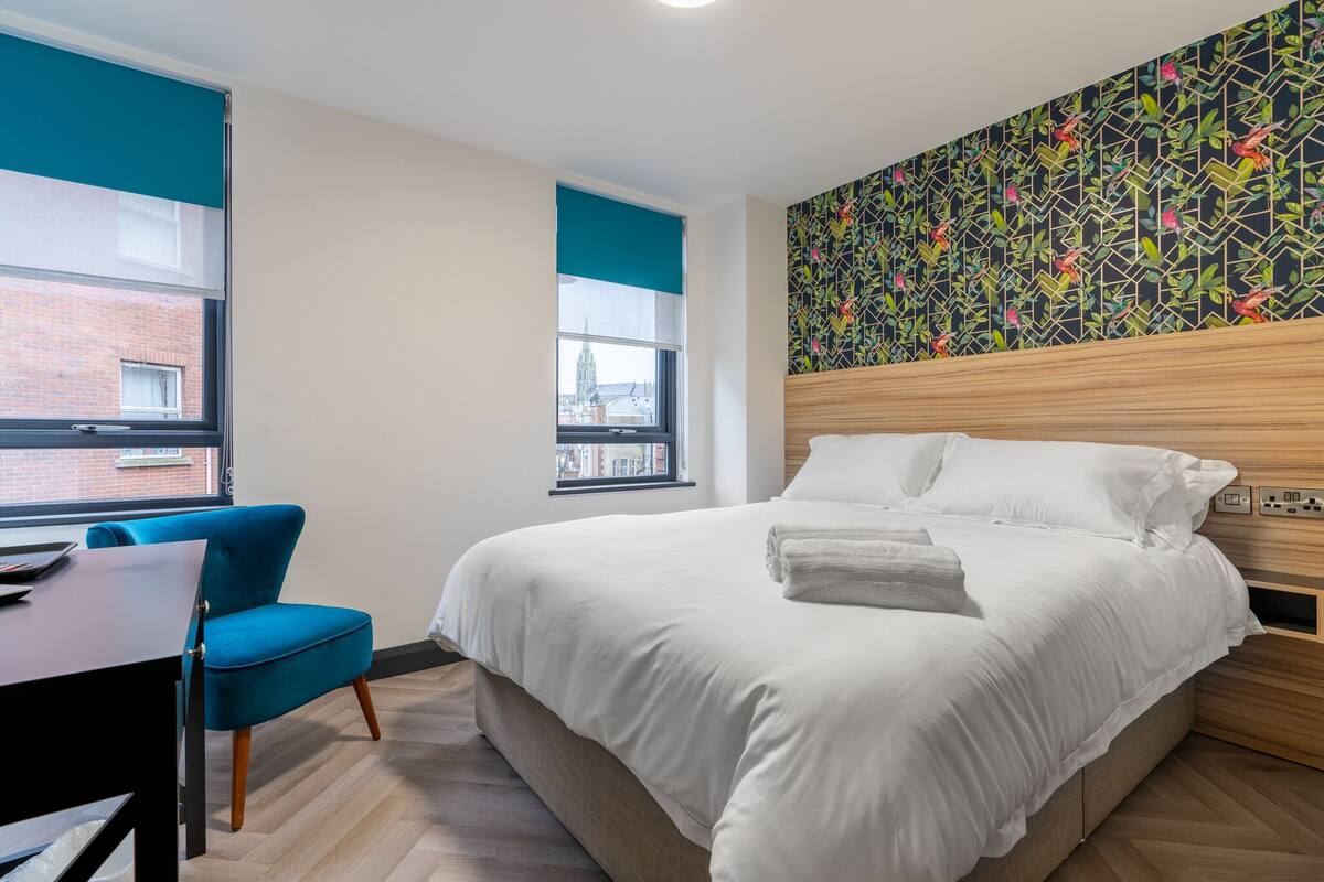 A hotel room with a bed, a desk, a chair, and a window with a view of a building.