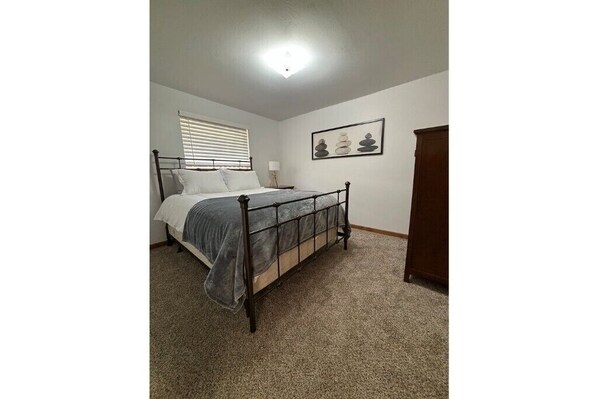 Une chambre à coucher avec un lit, une fenêtre avec des stores, une table de chevet avec une lampe et un tableau encadré au mur.