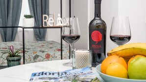 Un tavolo con una bottiglia di vino rosso, due bicchieri da vino, una ciotola di frutta, una candela e una rivista.