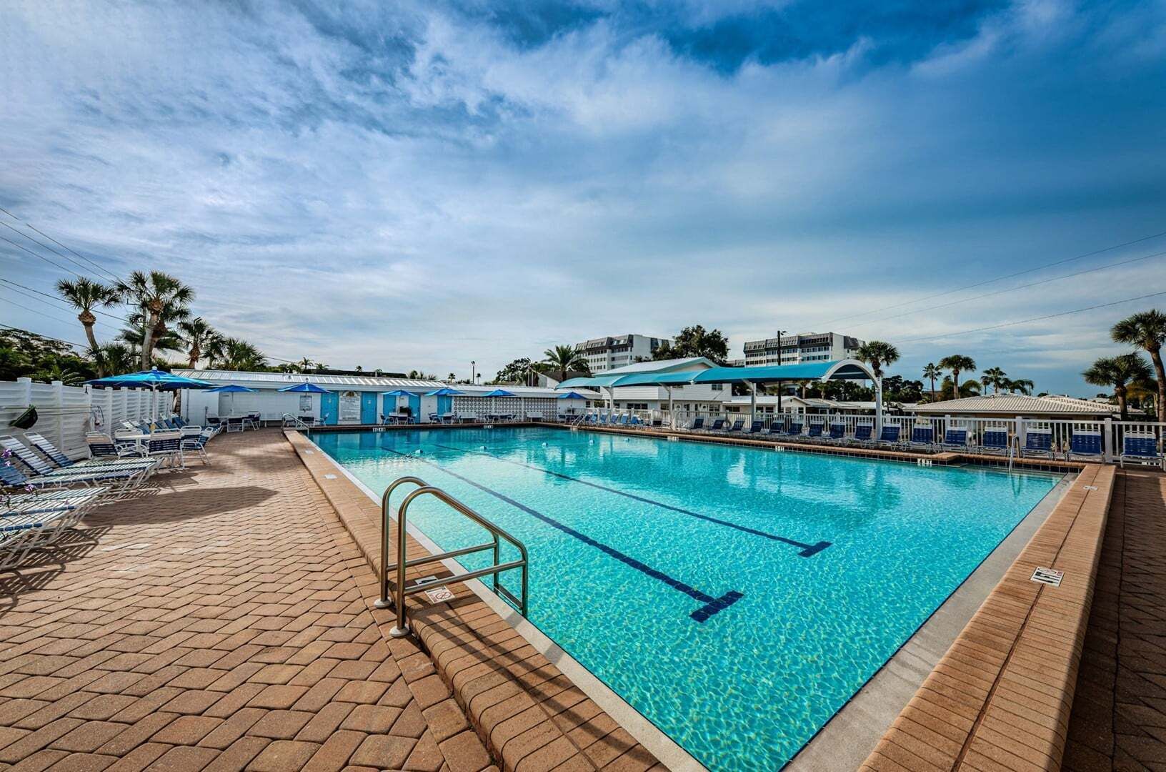 A swimming pool with lounge chairs and umbrellas, surrounded by a white fence and palm trees.