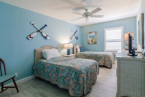 A bedroom with two beds, a ceiling fan, a window with blinds, and a desk with a lamp.