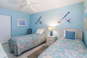 A bedroom with two beds, a ceiling fan, a nightstand with a lamp, and wall decorations including a framed picture and two paddleboards.