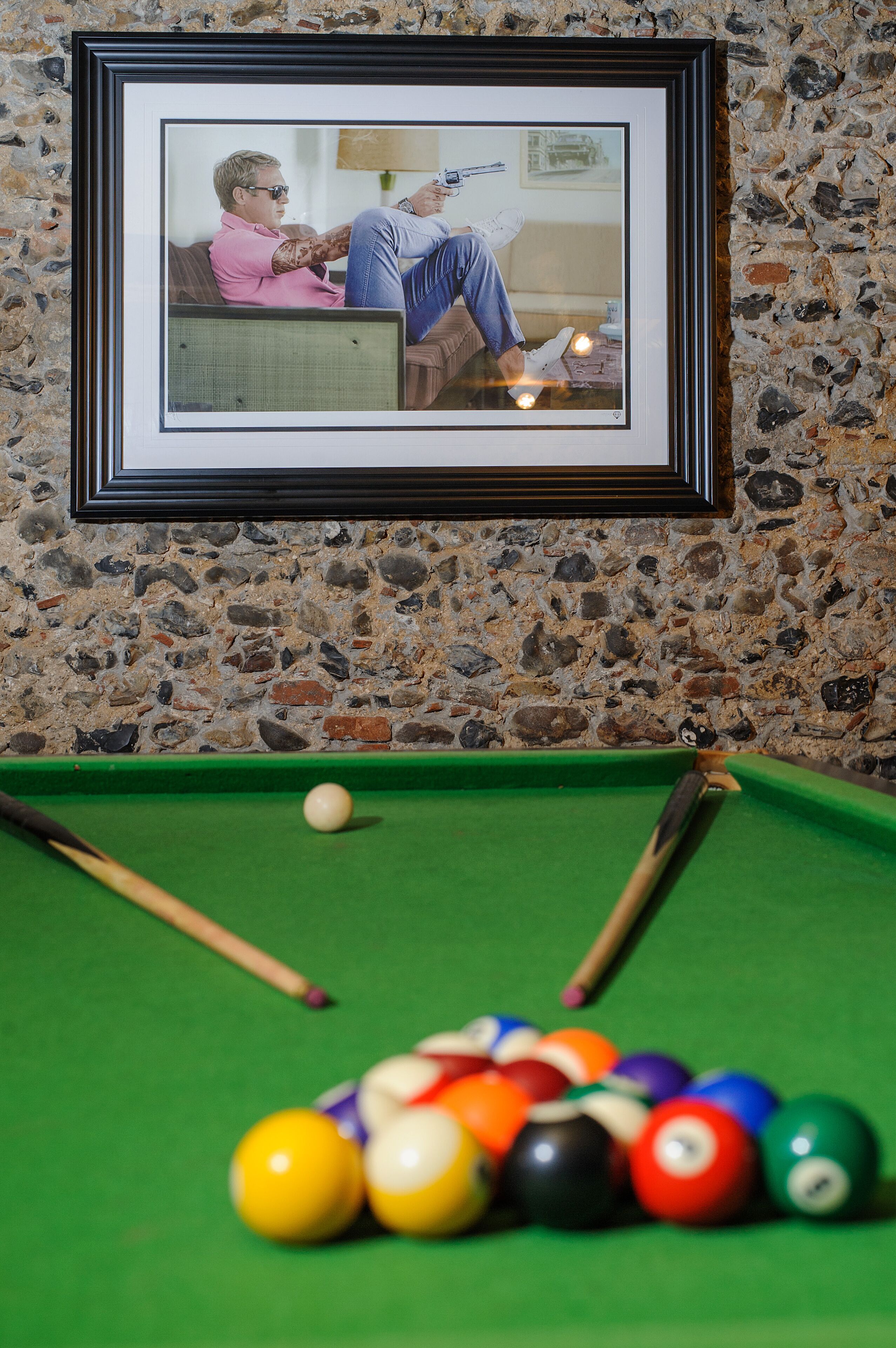 Une photographie encadrée représentant une personne se relaxant sur un canapé, une table de billard avec des billes, et un mur en pierre.