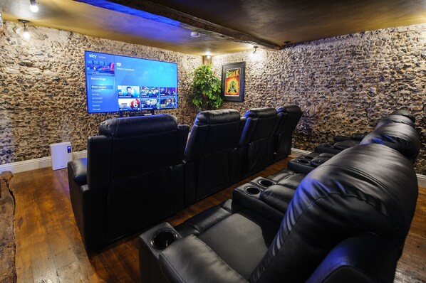 A home theater with leather reclining chairs, a large flat-screen TV, and a stone wall. - Luxury 2 Bed Manor House Hot Tub Cinema Room Skytv (Saffron Walden)